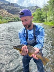 Flyfishing success: Man holding a brown trout in a river, mountains and trees in background. Flyfishing in Boise, Idaho.