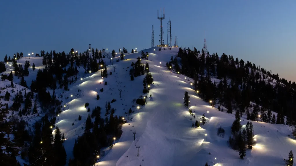 Night ski resort view with illuminated slopes, trees, and communication towers against a dark blue sky. Winter night operations.