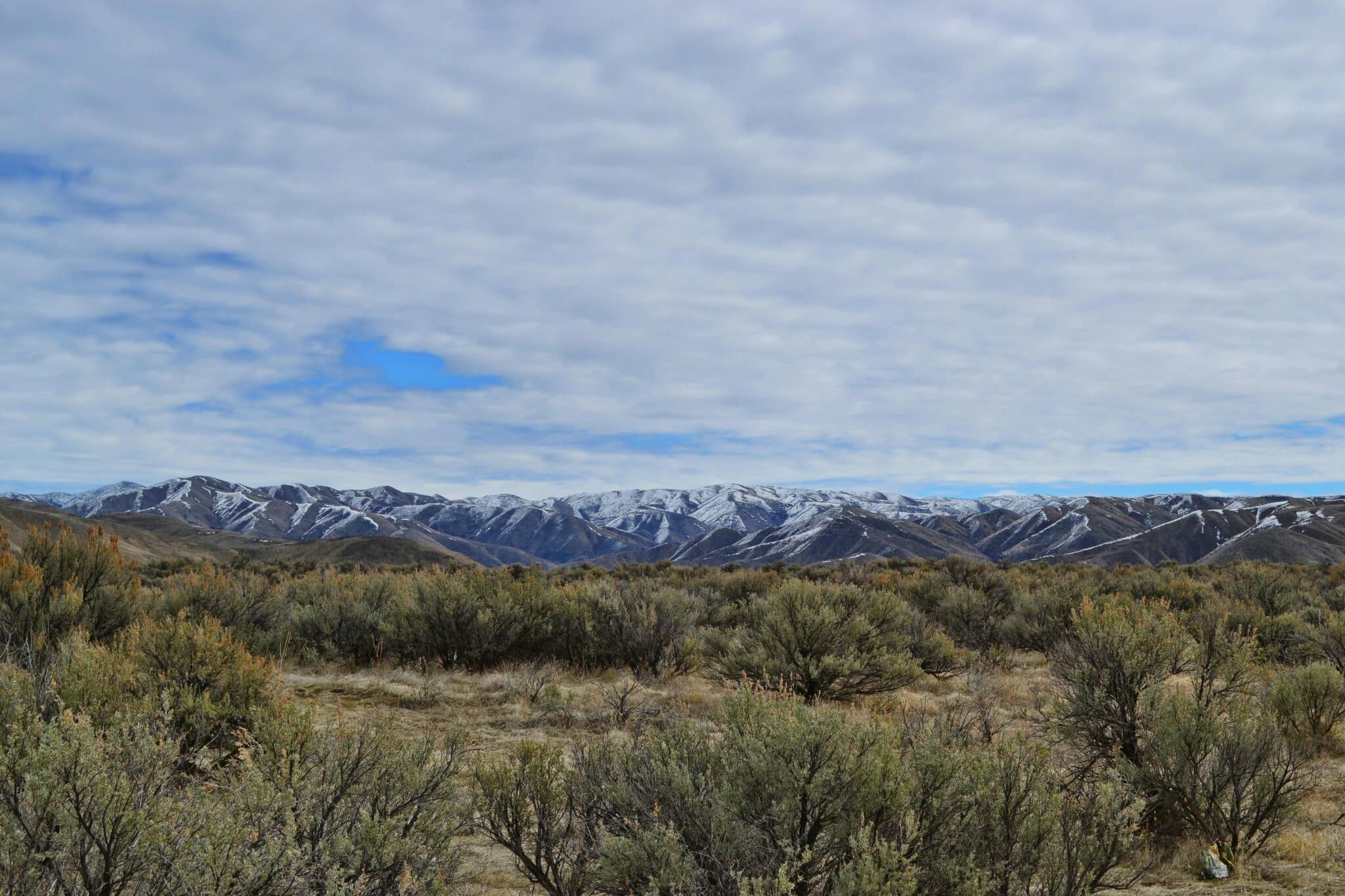 Wide view of the north end of Boise with sagebrush fields in the foreground, snow-dusted mountains in the distance, and a cloudy blue sky.